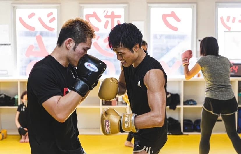 黄色マットのジム内で、黒いウェアとグローブを着けた2人の男性会員が笑顔で向かい合いスパーリングをする様子