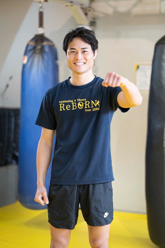 黄色マットのジムで、両脇に青と黒のサンドバッグがある空間で、ReBORNロゴ入りの黒いTシャツ姿に右手のパンチを突き出し笑う池田幸司(現役チャンピオン)