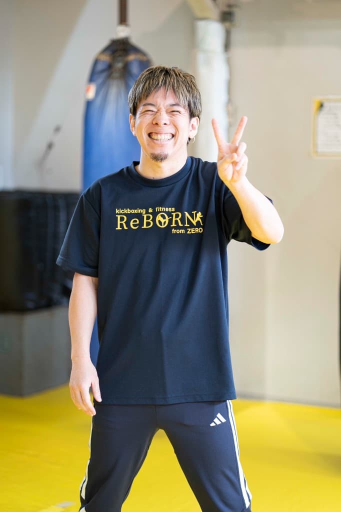 黄色マットのジム内で、ReBORNロゴ入りのネイビーTシャツとトラックスーツ姿で左手をピースにして笑顔を見せる正野宗一郎(プロキックボクサー)